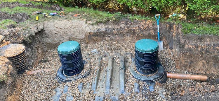 A partially dug trench reveals two large underground drainage systems with green covers. Gravel surrounds the structures, and a shovel is stuck in the ground nearby. A drainage contractor might be needed as pipes extend from each unit into the soil, hinting at potential blocked drains.