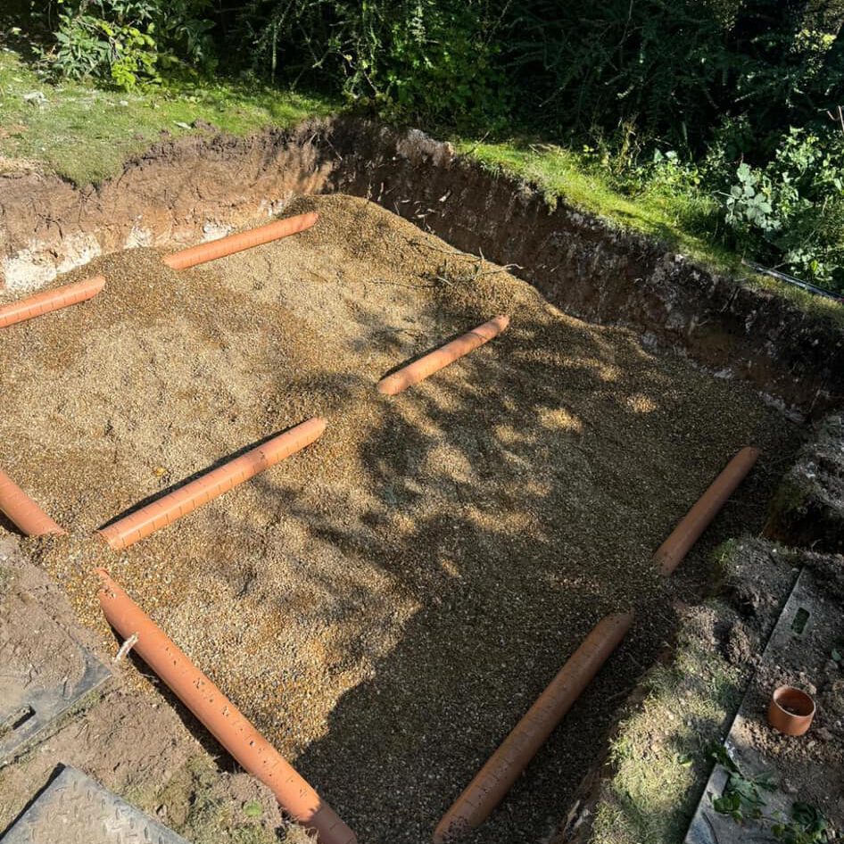 A rectangular hole in the ground filled with sand and gravel, featuring several evenly spaced orange drainage pipes, serves as an effective soakaway system. The surrounding area is grassy with some bushes.