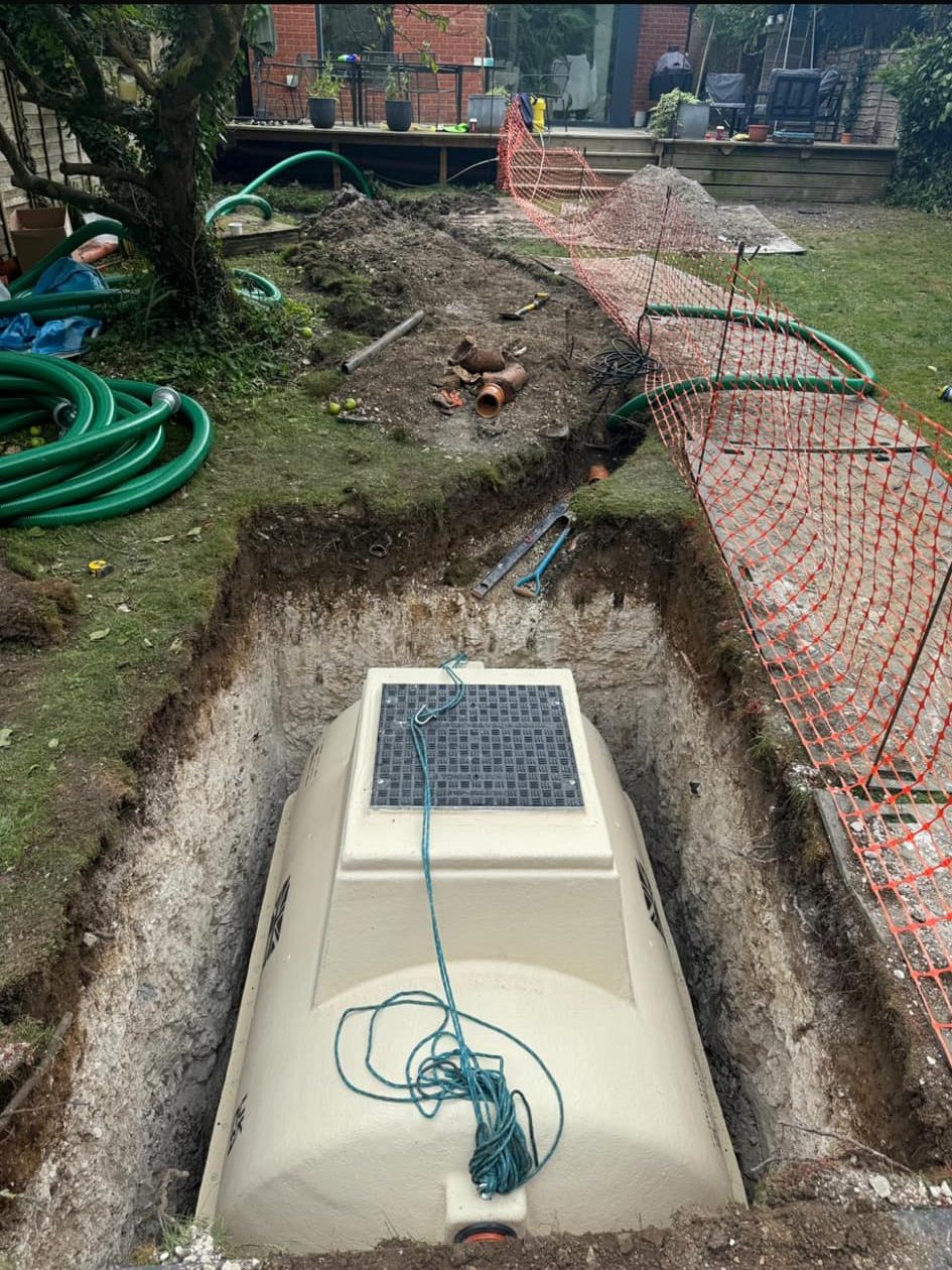 A partially buried tank with a grille on top is installed in an excavated pit, expertly handled by a drainage contractor, in a garden. A blue cable is attached to it. Green hoses, orange safety netting, and various tools are scattered around, with a brick wall and plants in the background.