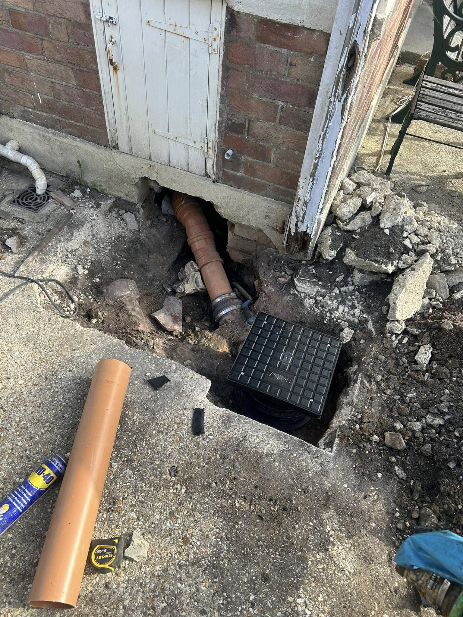 A construction site reveals a damaged wall with an exposed brown pipe leading into the ground. A black utility box sits in a small pit of broken concrete and bricks, indicating blocked drains. Nearby lies a detached pipe segment and a blue tool on the ground.