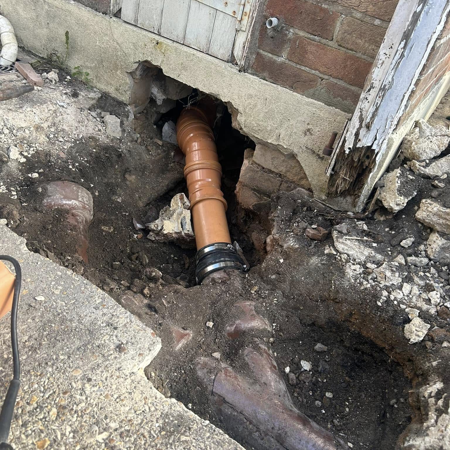 A damaged brick wall and foundation reveal an orange-brown plumbing pipe, indicating recent repair work by a drainage contractor. Nearby, soil and concrete debris suggest excavation, possibly to address blocked drains or install soakaways for improved water management.