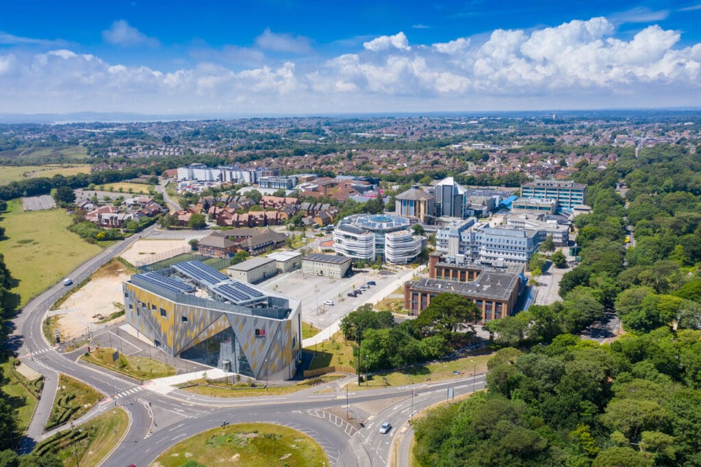 Aerial view of a campus with modern buildings, including a geometric-patterned structure. The campus is surrounded by trees and suburban housing, under a clear blue sky. Nearby, roads lead to facilities where CCTV surveys ensure well-maintained infrastructure in harmony with nature.
