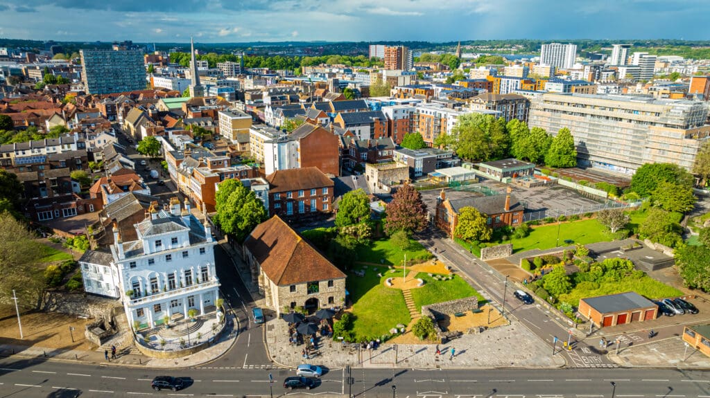 Aerial view of a cityscape featuring a mix of historic and modern buildings. In the foreground, a green park with pathways and trees seamlessly integrates soakwaways for effective drainage. Roads surround the area with some cars and people visible, while the background showcases dense urban development.