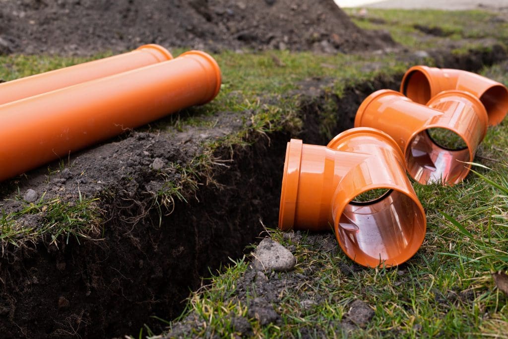 Several orange PVC drainage pipes and connectors are placed next to a freshly dug trench in a grassy area, indicating ongoing work by a drainage contractor. This setup suggests plumbing or sewer improvements, potentially involving soakaways or sewage treatment plants.