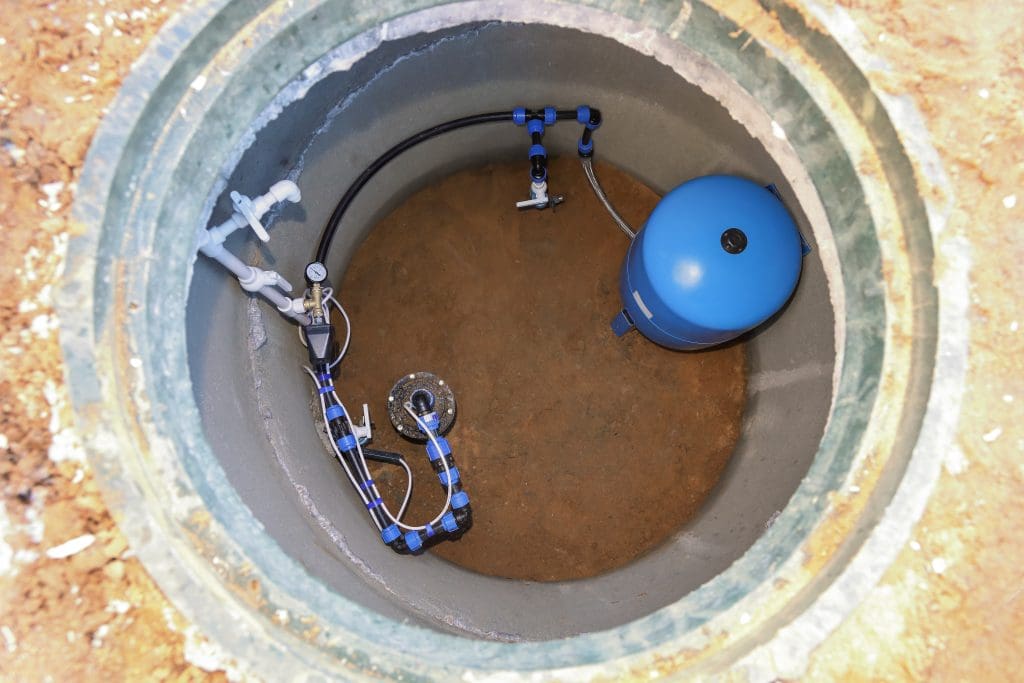 View of an open underground water well with a blue pressure tank and connected piping inside a concrete lining. The circular top edge and surrounding earth are visible, reminiscent of the meticulous work often seen by a skilled drainage contractor.