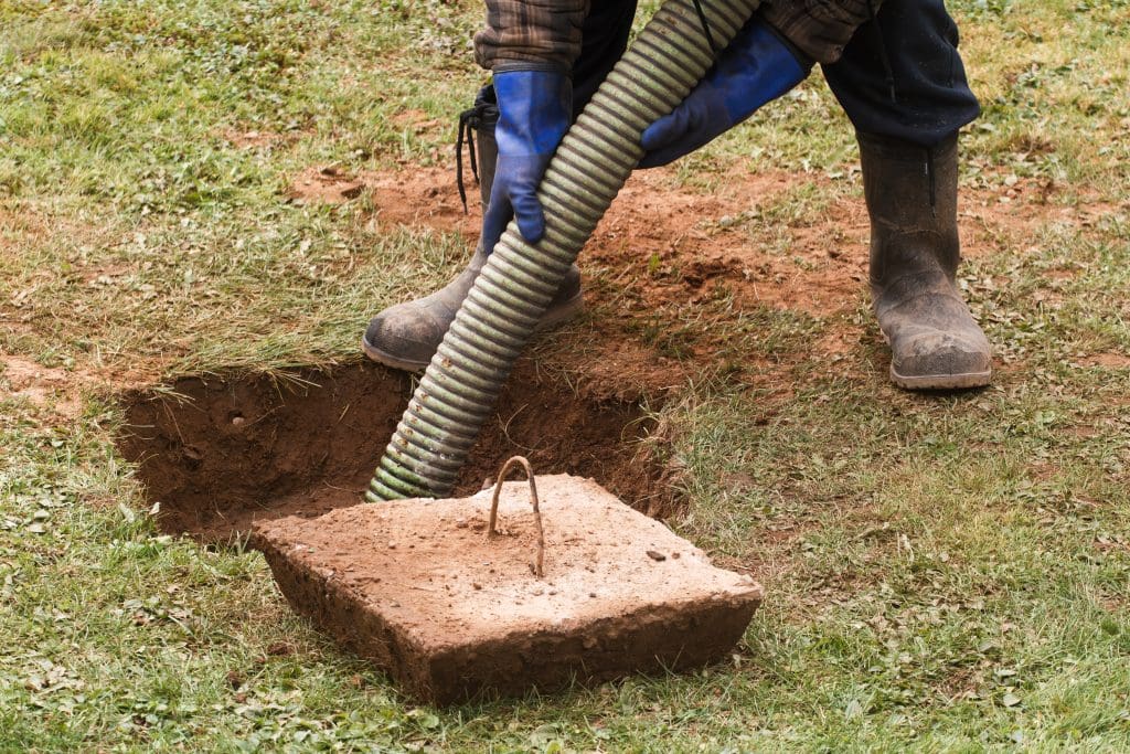 A person wearing blue gloves and boots is holding a green hose, operating near an open septic tank in a grassy area, perhaps addressing blocked drains. A removed concrete lid with a handle lies nearby, revealing the dug-up ground beneath.
