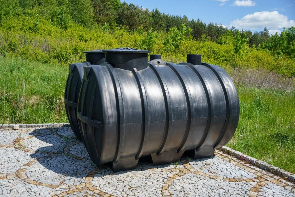 A large black plastic septic tank sits on a cobblestone platform amid a grassy area, designed with effective soakeaways in mind. In the background, there are trees and a blue sky with a few clouds.