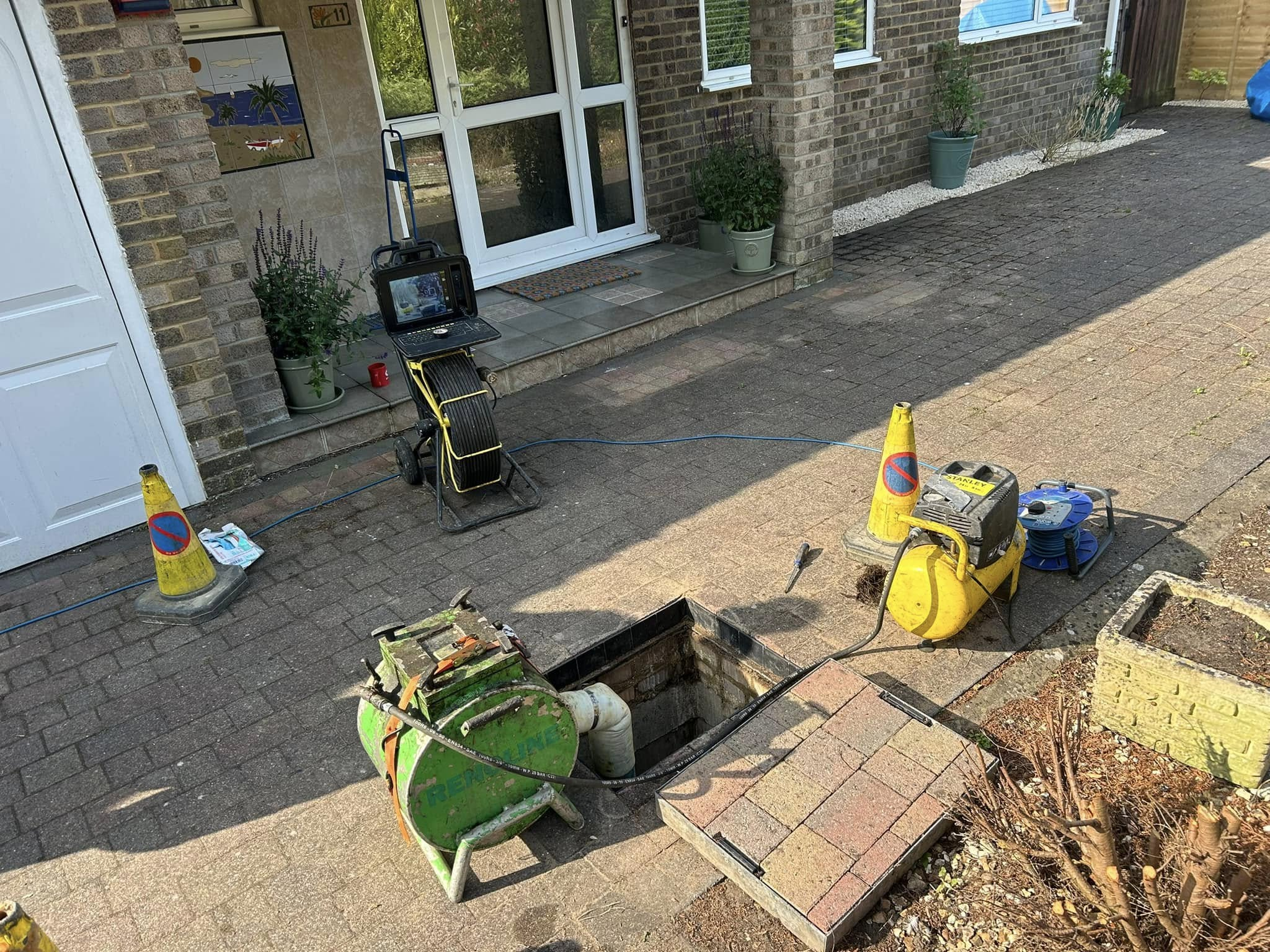 A driveway with an open manhole and traffic cones, featuring equipment for CCTV surveys, including a cable reel and monitor. This setup suggests maintenance work on blocked drains. A brick house entrance is visible in the background.