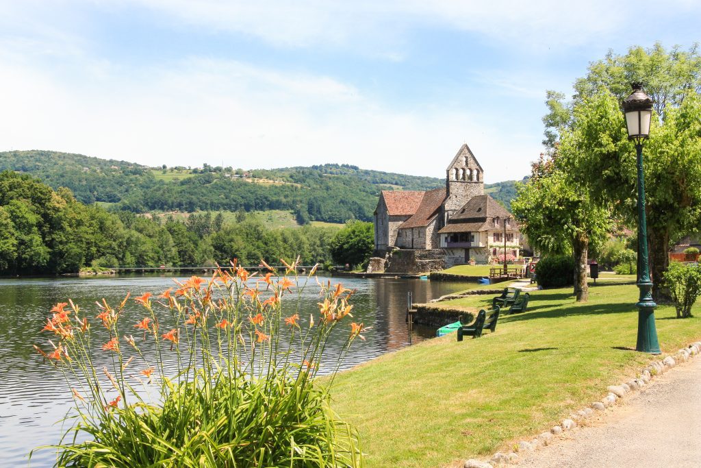 A stone church with a bell tower stands by a calm river, surrounded by green hills and trees. Orange flowers bloom in the foreground, and a lamppost and benches line the grassy riverbank under a blue sky.