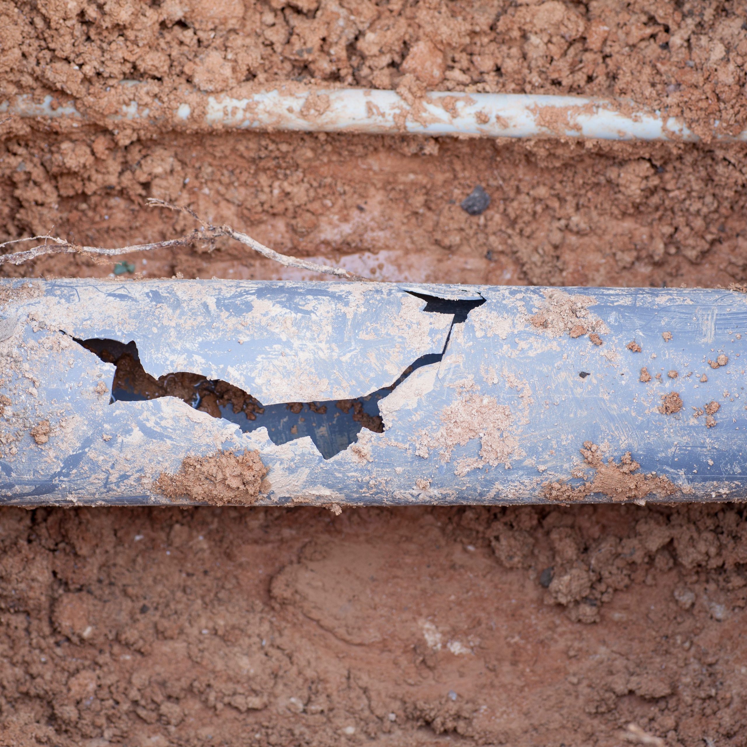 A cracked and damaged grey pipe is partially buried in dirt, its surface showing significant tear from a potential leak. The brown, loose soil suggests recent digging. Such conditions may warrant further investigation through CCTV surveys to assess the extent of the damage properly.