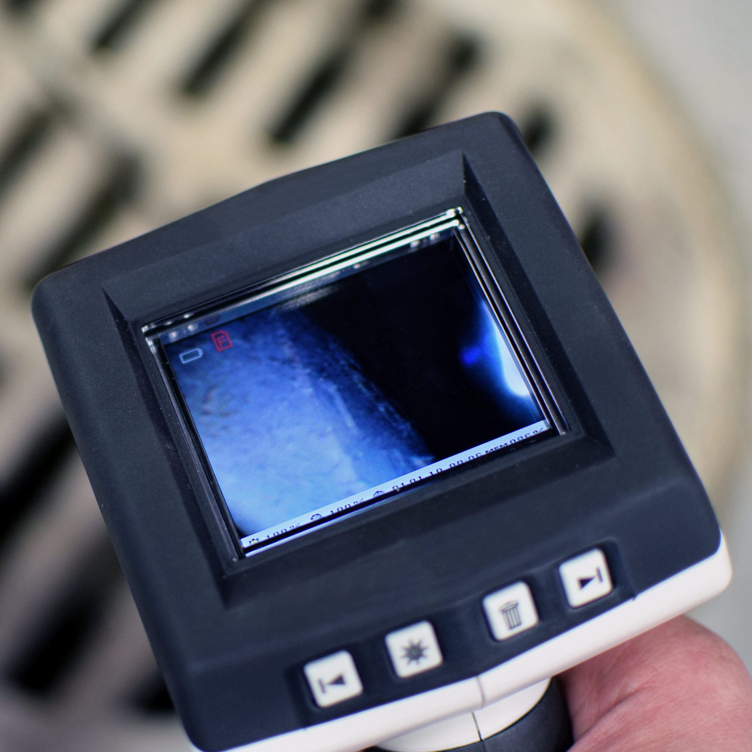 A person holds an inspection camera near a round metal grate with parallel bars, likely checking for blocked drains. The camera's screen displays a blue and gray-toned image against the concrete surface, typical of a drainage contractor at work.