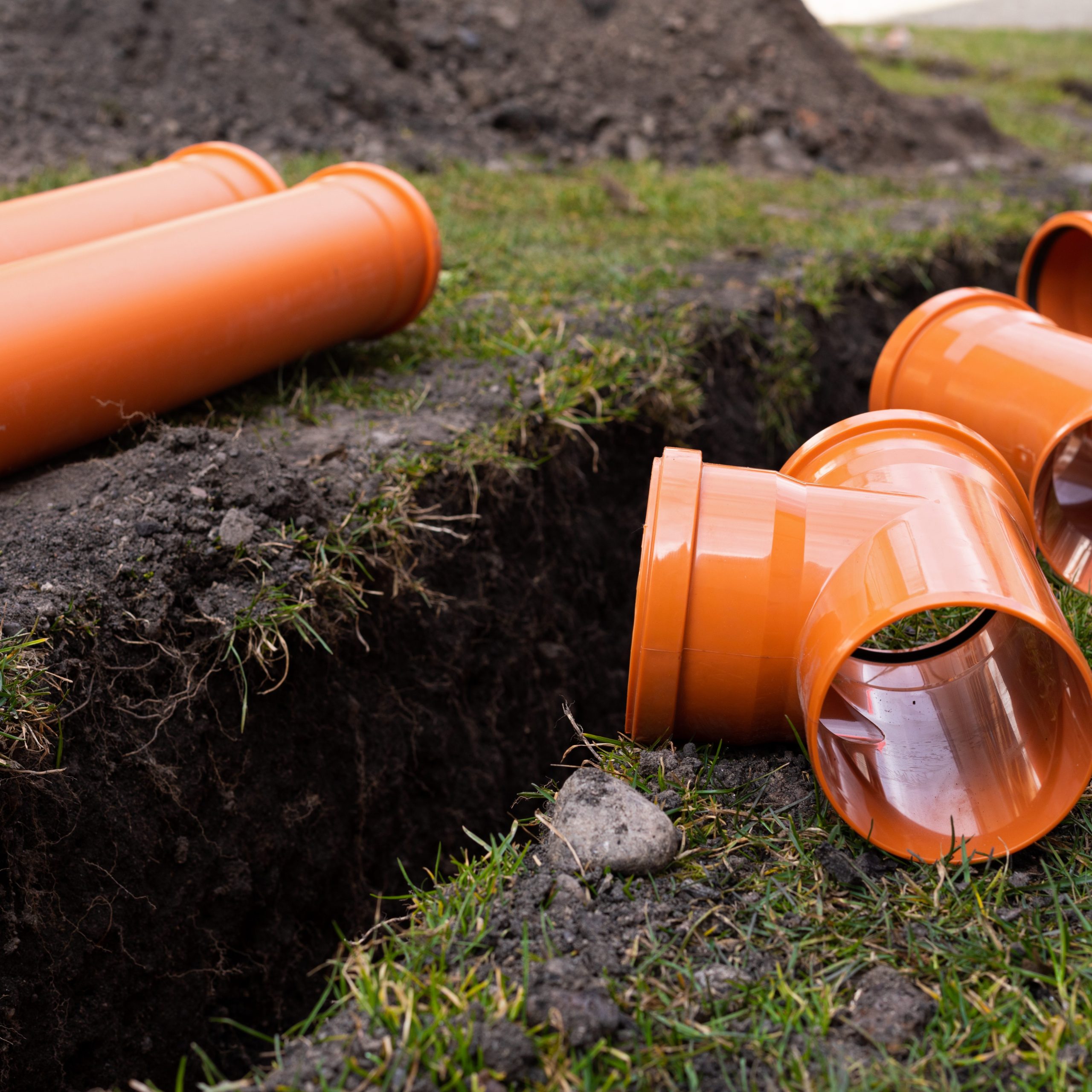 Several orange PVC drainage pipes and connectors are placed next to a freshly dug trench in a grassy area, indicating ongoing work by a drainage contractor. This setup suggests plumbing or sewer improvements, potentially involving soakaways or sewage treatment plants.