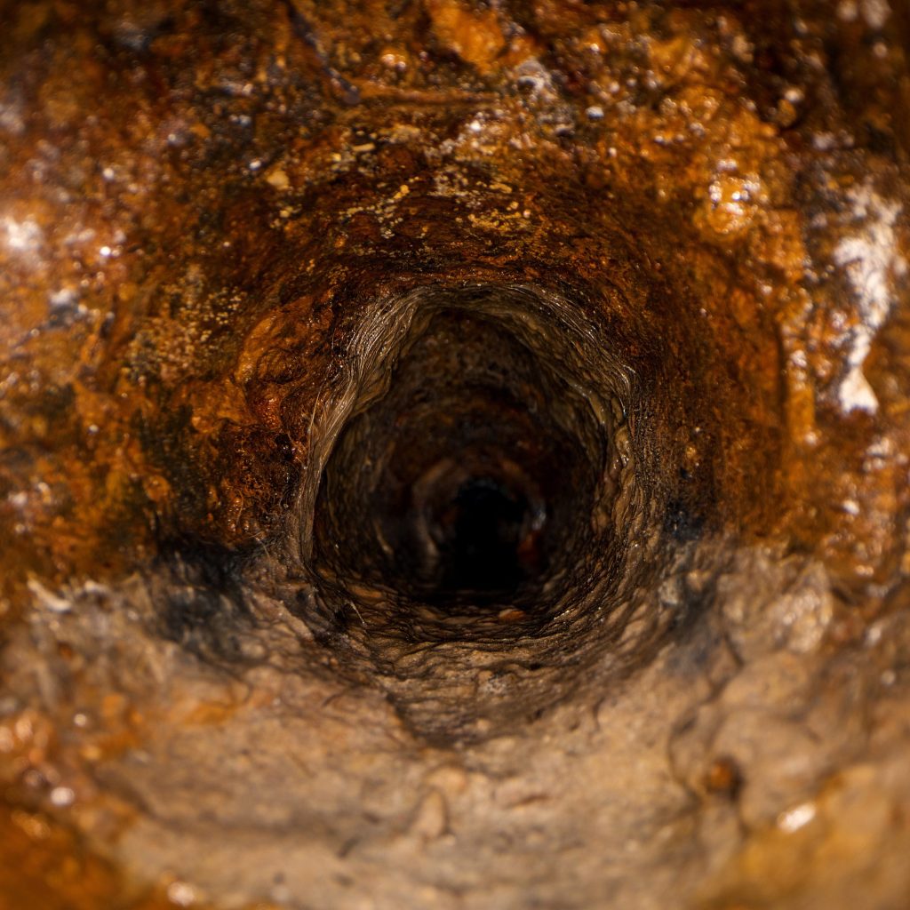 Close-up of a corroded metal pipe interior, typical in drain re-lining projects. The rough, rust-covered walls create a textured appearance, while the dark, narrow passage fades to black towards the center, emphasizing depth.