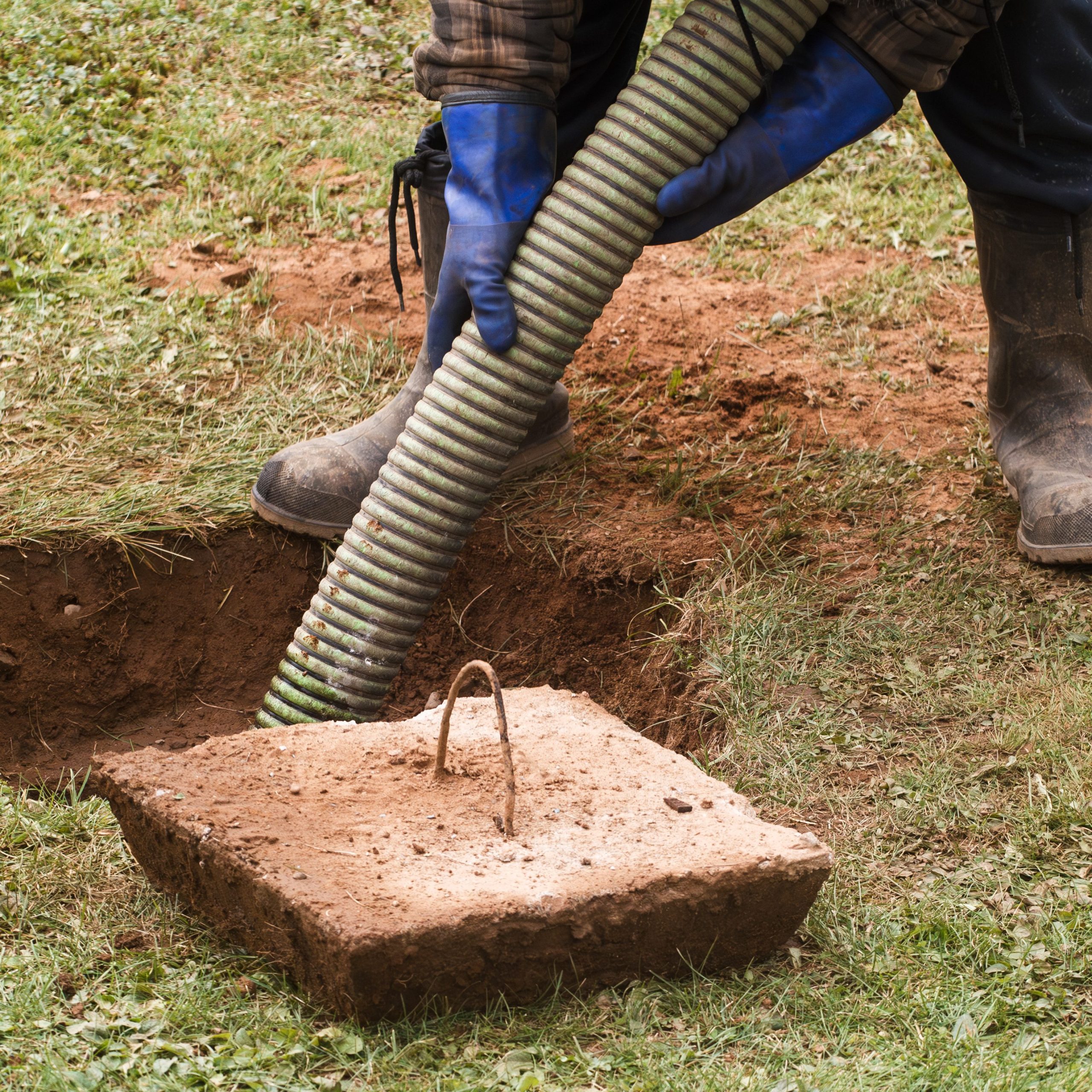 A person wearing blue gloves and boots is holding a green hose, operating near an open septic tank in a grassy area, perhaps addressing blocked drains. A removed concrete lid with a handle lies nearby, revealing the dug-up ground beneath.