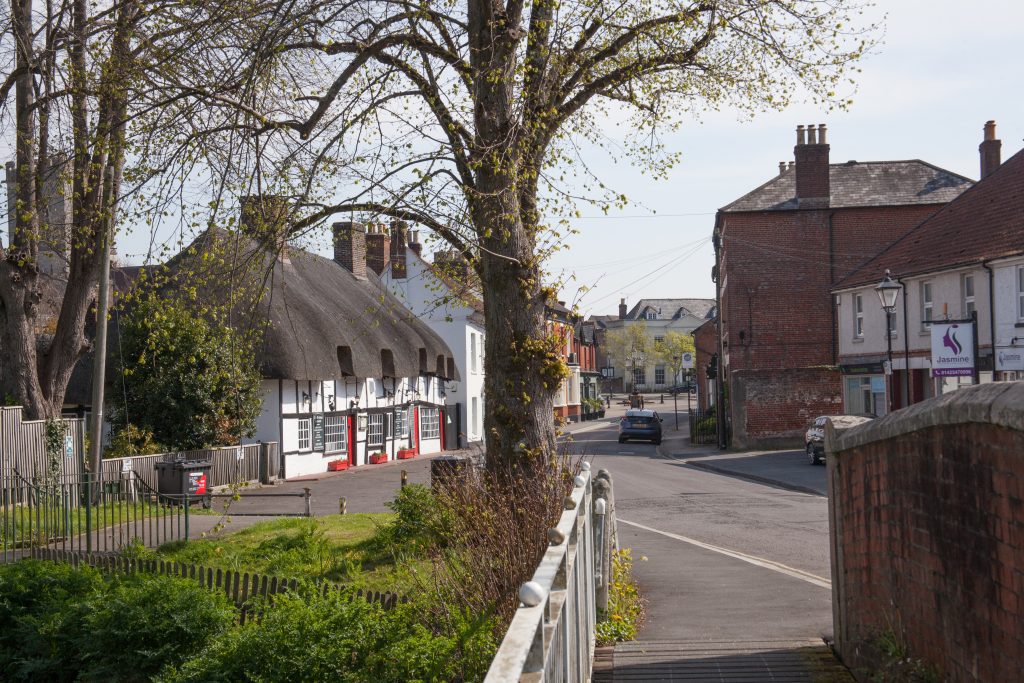 A quiet street in a small town features a white thatched-roof cottage, red-brick buildings, a tree with early spring leaves, and a sidewalk bordered by metal railings under a clear sky.