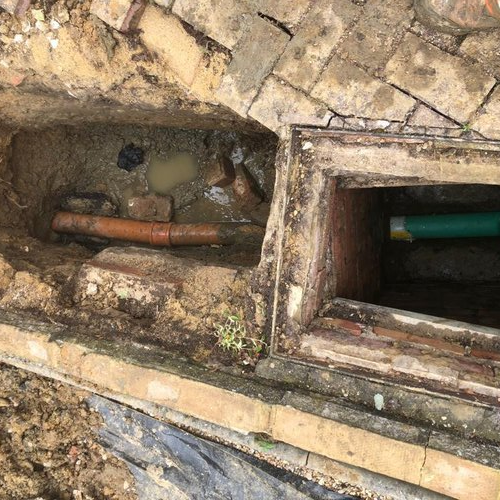 A construction site with two rectangular excavations in the ground. One pit reveals an orange pipe amidst muddy soil, possibly needing a CCTV survey, while the other shows a green pipe, surrounded by bricks and soil. There's a section of brick paving at the top.