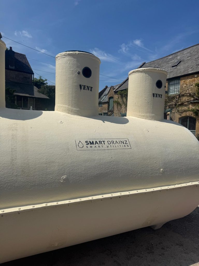 A large, cream-colored cylindrical tank labeled "SMART DRAINZ" with two vertical vents stands outdoors under a blue sky, with stone buildings visible in the background.