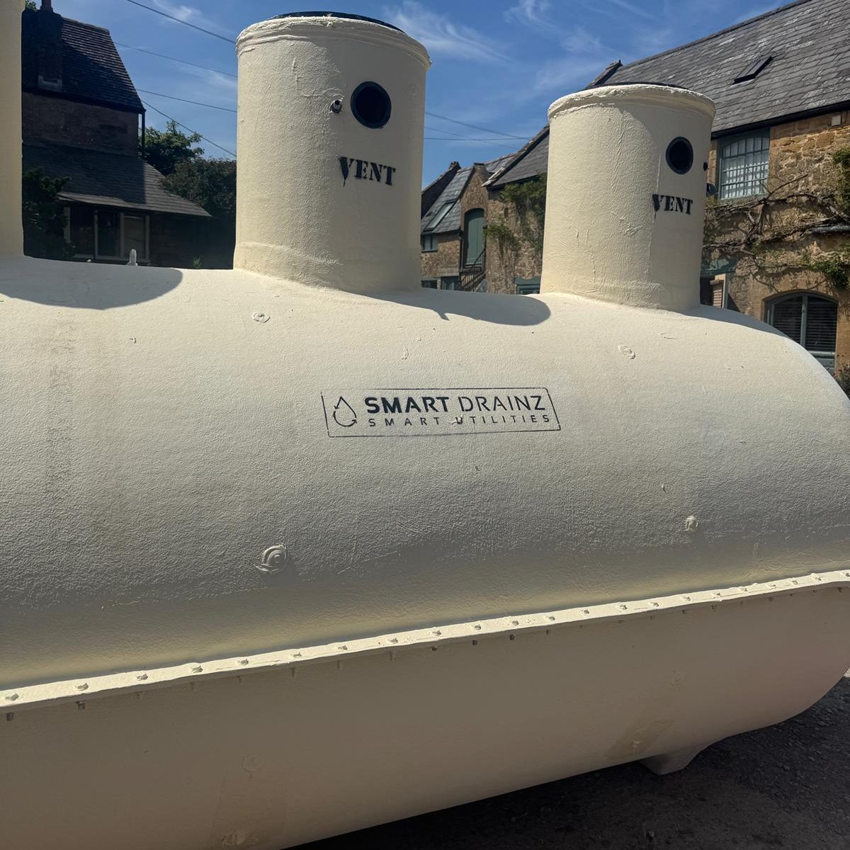 A large, cream-colored cylindrical tank labeled "SMART DRAINZ" with two vertical vents stands outdoors under a blue sky, with stone buildings visible in the background.