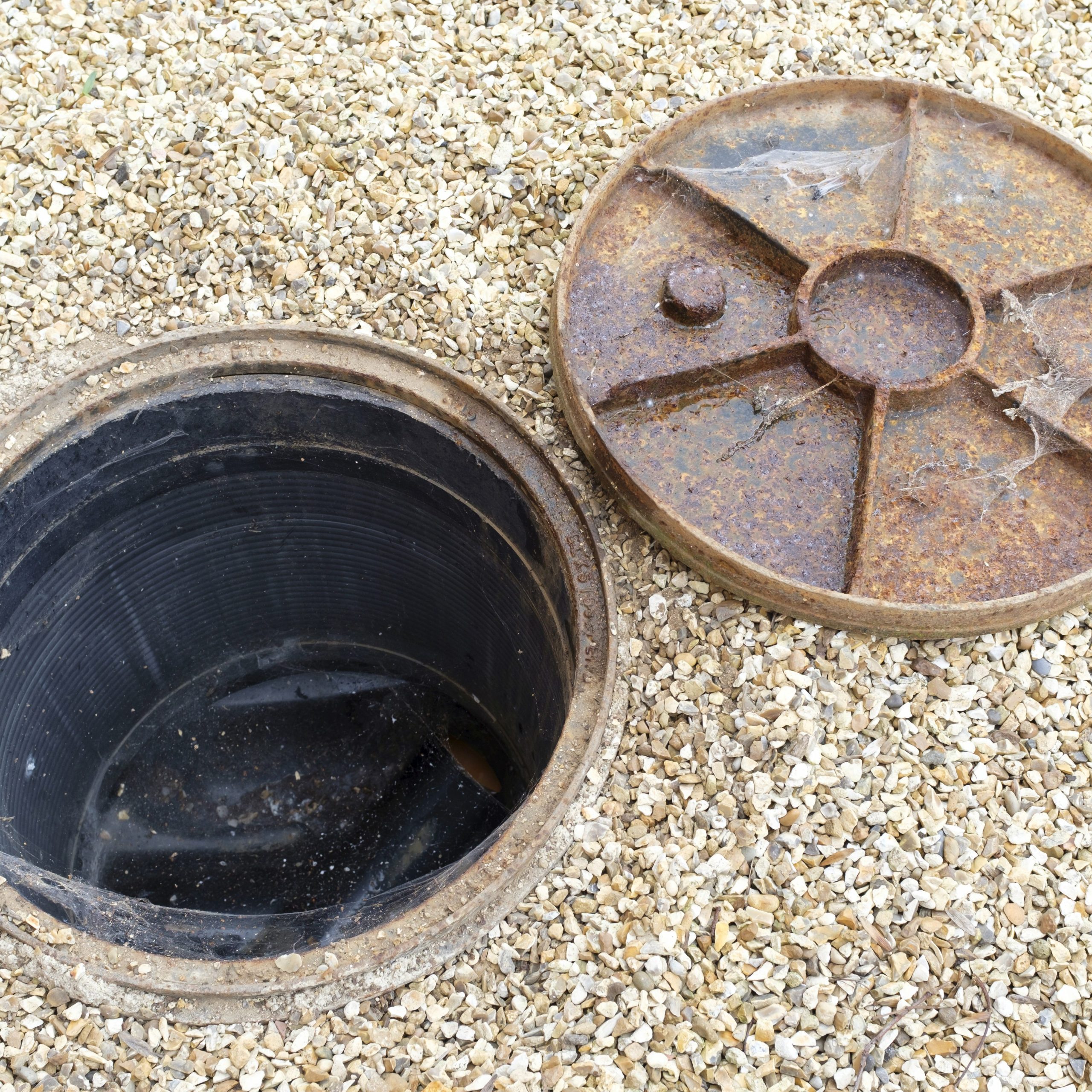 A circular, open manhole on a gravel surface with a rusted metal cover lying beside it, reminiscent of the entrances used in underground CCTV surveys.
