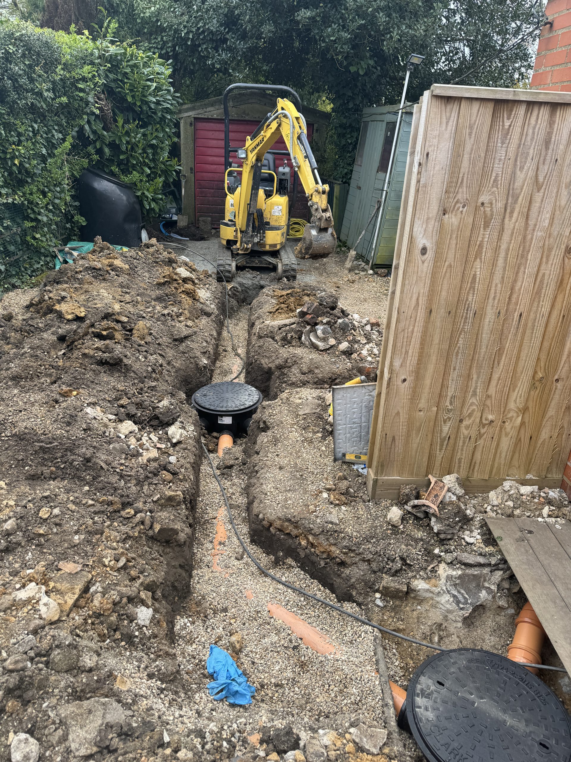 A small yellow excavator sits in a backyard, next to a trench with exposed orange drainage pipes and inspection covers. Soil and gravel are piled along the edges, with a wooden fence and shed nearby.