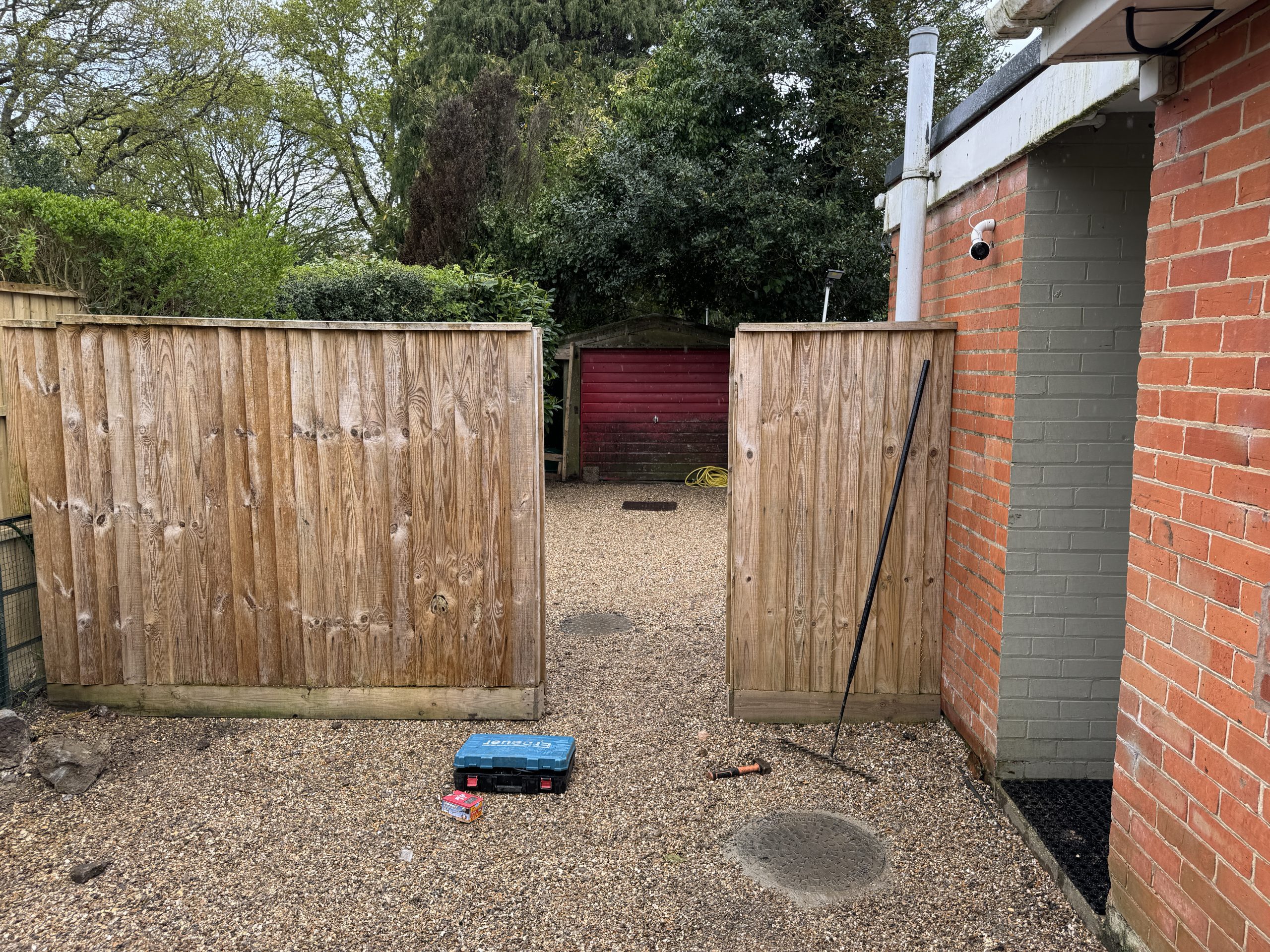 A gravel-covered backyard with a wooden fence gate open in the middle, a red wooden shed at the back, and a brick building on the right. Tools and a blue tool box are on the ground near the gate.