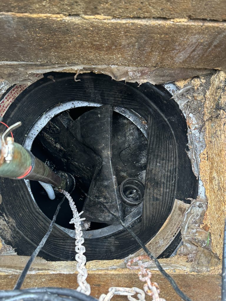 View looking down into a circular, industrial opening with pipes, chains, and dark, greasy machinery inside. The opening is surrounded by worn metal and debris.