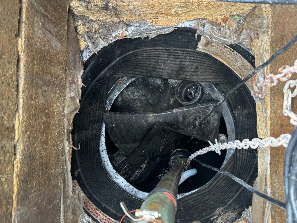 Close-up view of the inside of a large industrial pipe or manhole with metal chains, cables, and a cylindrical instrument or sensor extending into the dark, grimy interior.