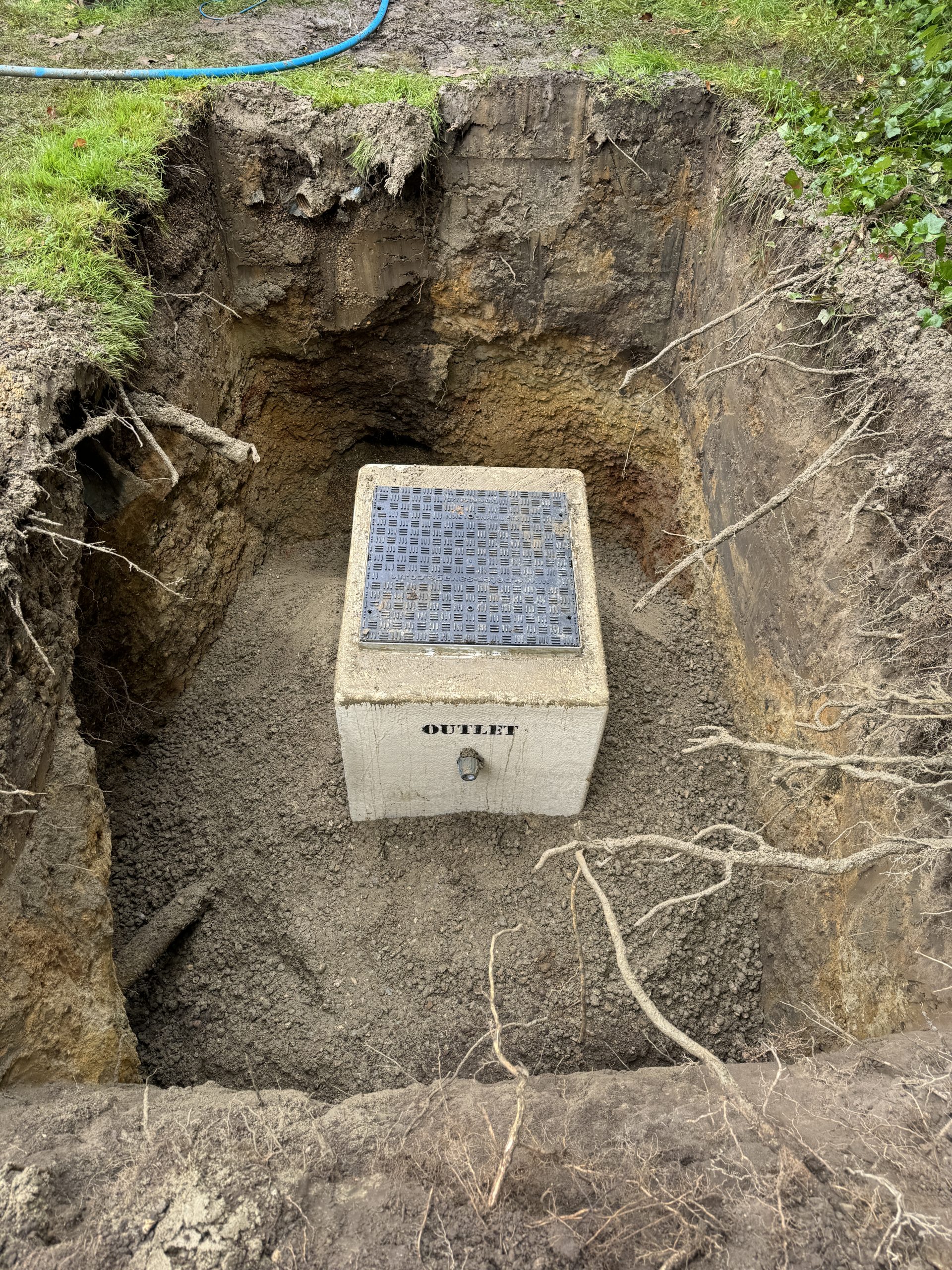 A concrete drainage box labeled "OUTLET" is installed in a deep, freshly dug rectangular pit, surrounded by soil layers and exposed roots. A blue hose lies on the grass at the top edge of the hole.