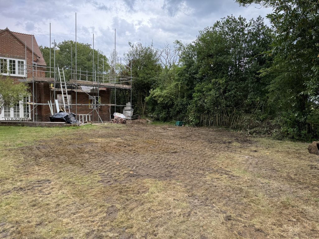 A patchy, muddy yard with tire tracks, next to a large house under construction with scaffolding. Piles of bricks and building materials are visible, and dense trees line the right side.