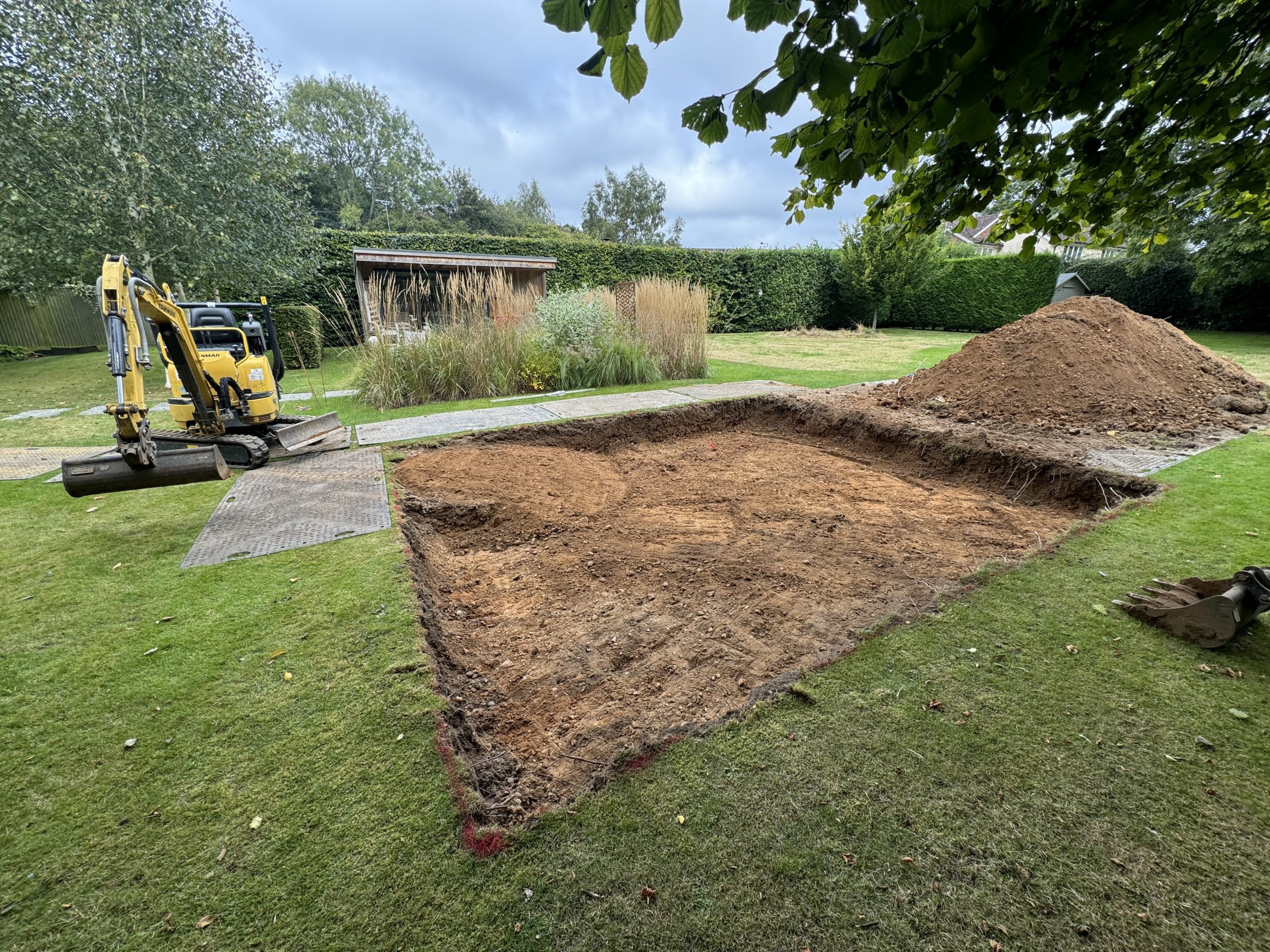 A rectangular patch of soil has been excavated in a grassy backyard. A mini excavator and a large pile of dirt are nearby. Bushes, trees, and a garden shed are in the background.