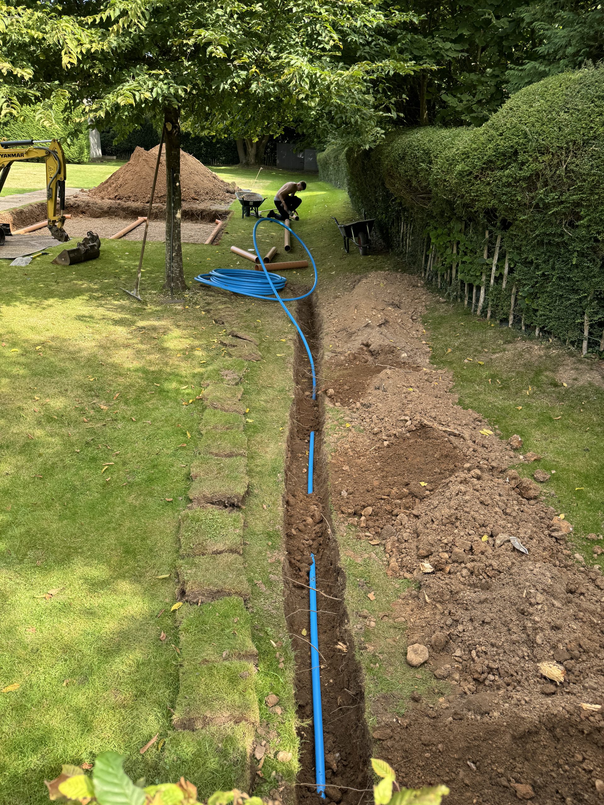 A person installs a blue pipe in a long, narrow trench dug through a grassy yard surrounded by trees and hedges. Construction equipment and soil piles are visible in the background.