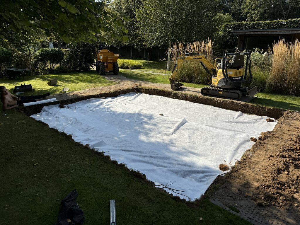 A rectangular patch of earth, lined with white fabric, is being prepared in a garden. A small yellow excavator and a dumper are parked nearby, surrounded by green grass and tall plants.