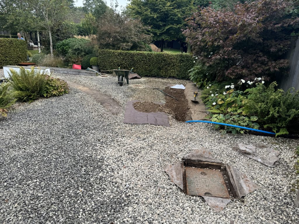 A garden area with gravel, plants, and bushes is under construction. A wheelbarrow, tools, and a blue hose are visible. Some soil is spread on a tarp, and a partially open utility cover is in the foreground.