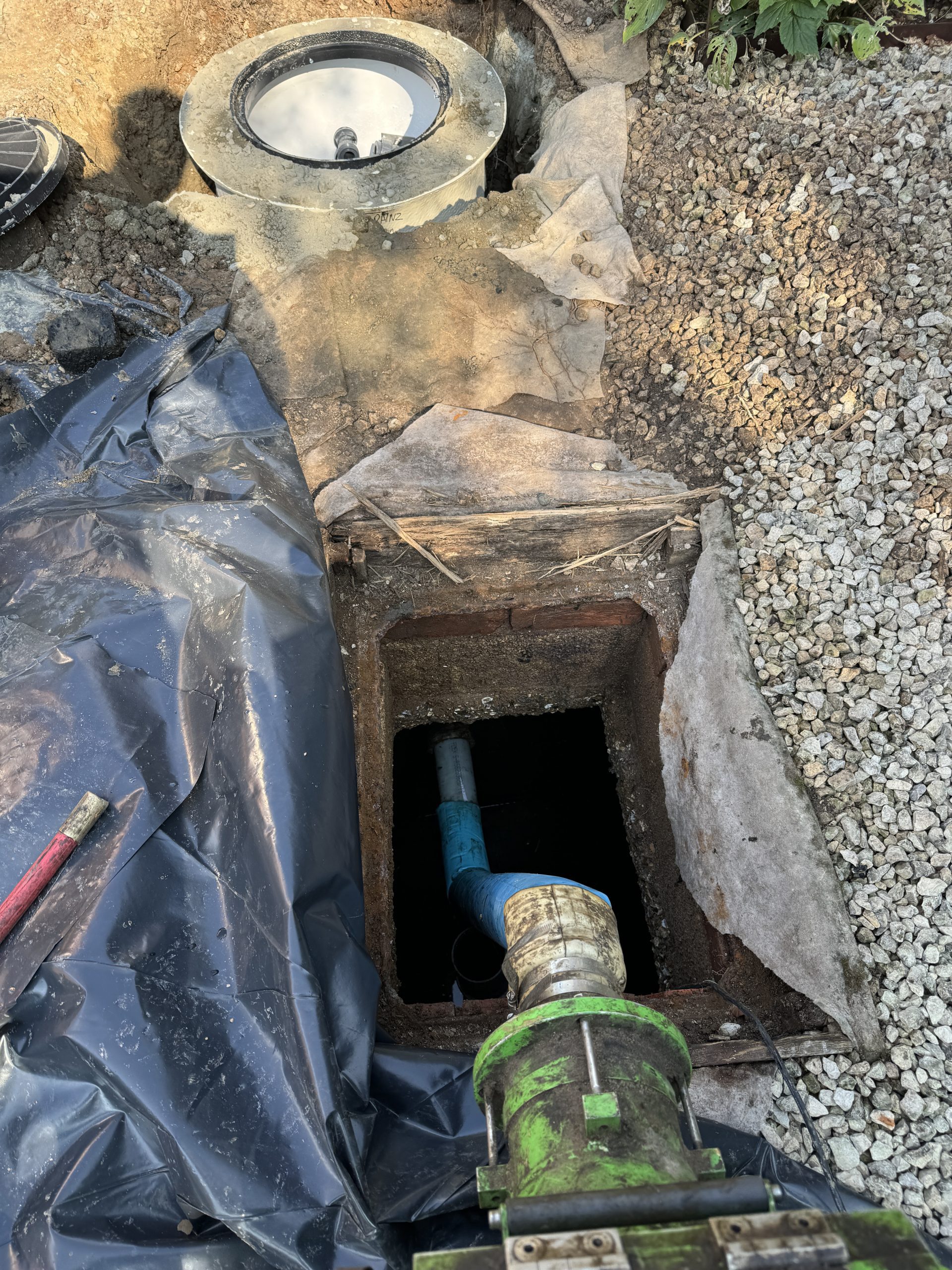 A rectangular hole in the ground with a green and blue pipe inside, surrounded by gravel, black plastic sheeting, and a round concrete cover nearby.