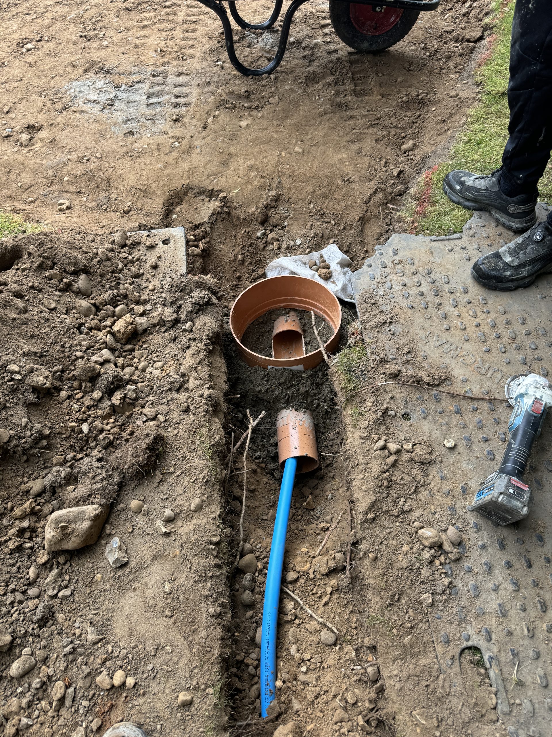 A shallow trench with exposed soil contains a blue pipe and a round brown drainage system. A person stands nearby with work boots, and tools, rocks, and a wheelbarrow are visible around the excavation site.