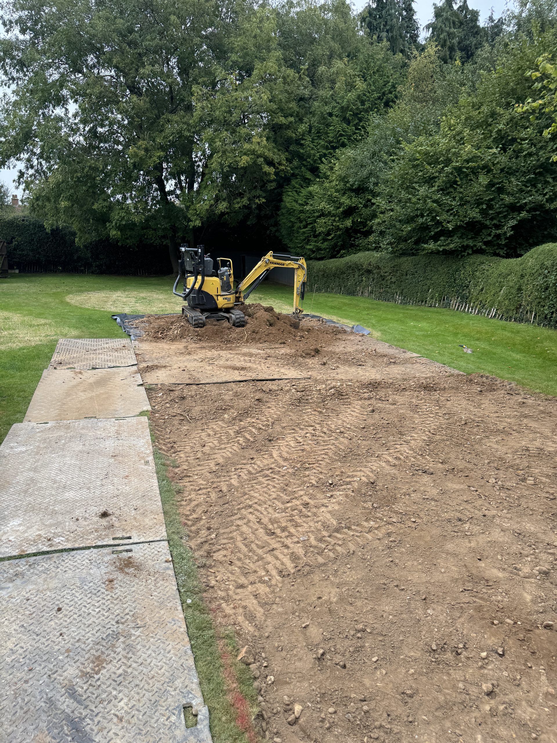 A small yellow excavator is digging up soil in a garden surrounded by grass and trees, with metal plates laid out on the ground in the foreground.