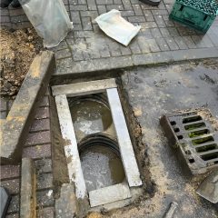 A sidewalk with an open rectangular manhole, some exposed pipes, construction tools, a traffic cone, a crate, and a plastic bag nearby. The surrounding area is wet and muddy.