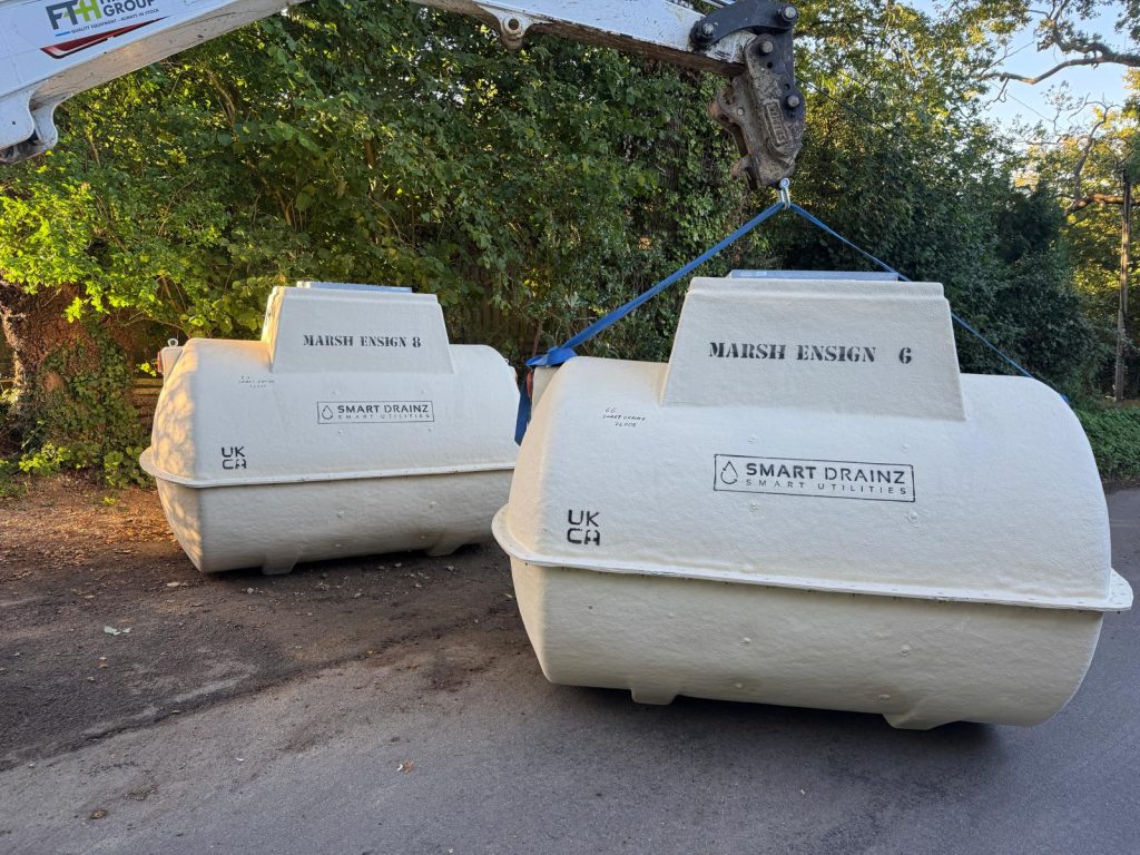 Two large white Marsh Ensign tanks labeled “SMART DRAINZ” are being moved by a crane on a rural road, with trees and greenery in the background.
