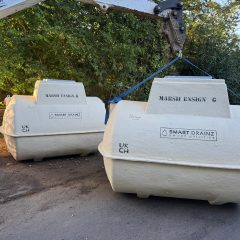 Two large white Marsh Ensign tanks labeled “SMART DRAINZ” are being moved by a crane on a rural road, with trees and greenery in the background.