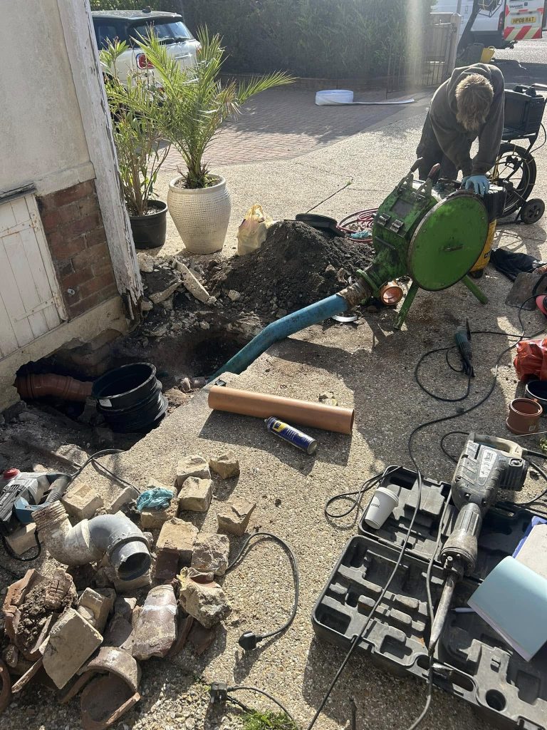 A worker operates machinery near a dug-up area by a house. Tools, pipes, and bricks are scattered on the ground, and a blue hose is connected to equipment. A white car and potted plant are visible in the background.