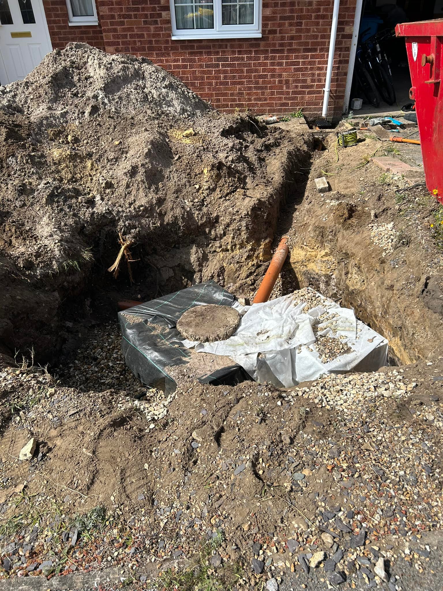 A large hole is dug in front of a brick house, exposing a buried black plastic tank and an orange pipe. Piles of dirt and gravel surround the excavation site, with some construction materials visible.