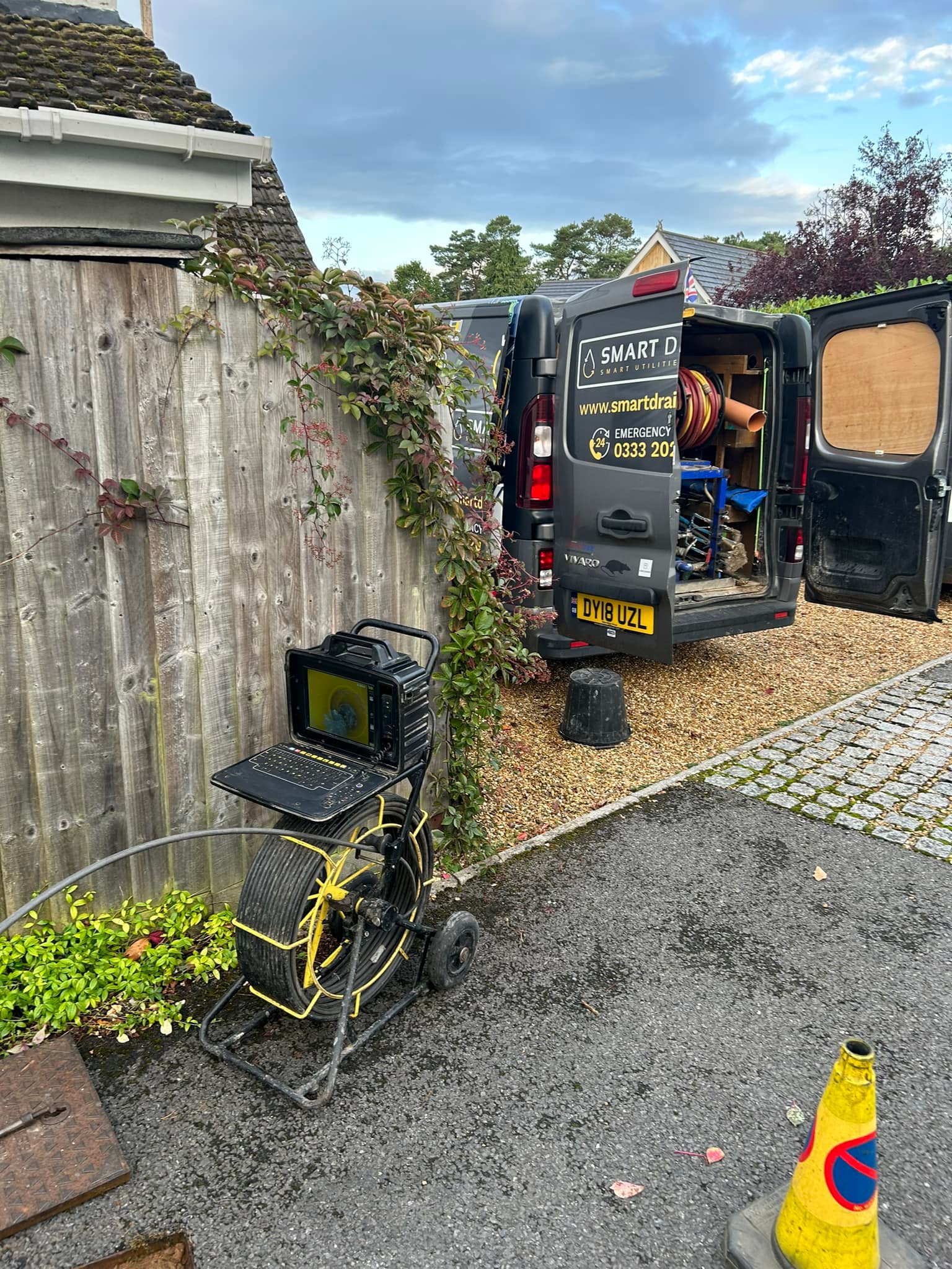 A black van with open rear doors is parked on a gravel driveway. In the foreground, a drain inspection camera and monitor are set up near a wooden fence, with a yellow traffic cone on the pavement.