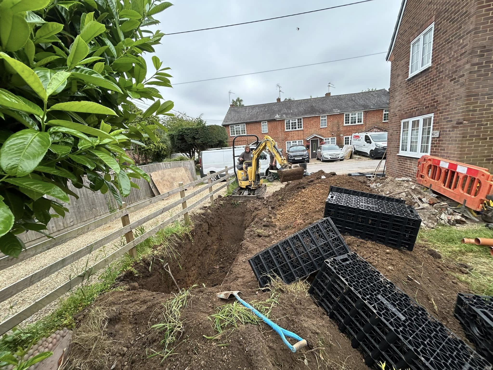 A small excavator digs a trench in a residential yard. Black plastic crates, soil piles, and tools are scattered nearby. Houses, parked cars, and a wooden fence are visible in the background under an overcast sky.