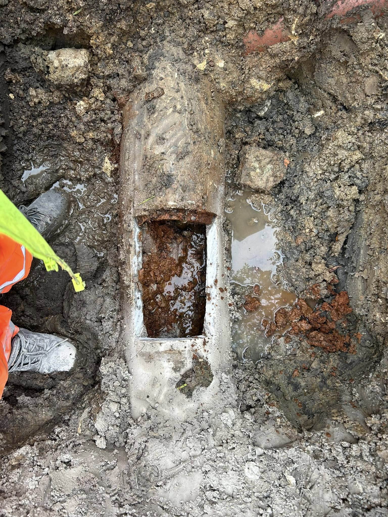 A large, exposed pipe buried in muddy soil with a rectangular section cut open, revealing rust and debris inside. A person in orange safety gear and boots stands nearby.