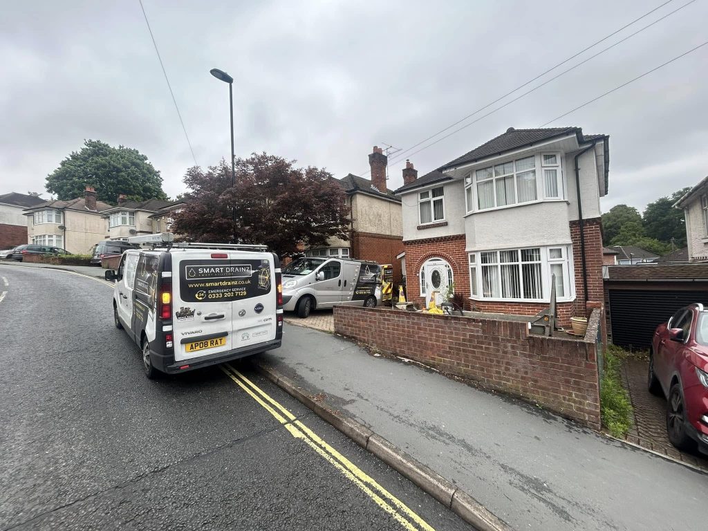 A white van with business signage is parked on a residential street outside a two-story brick house with a white door. Another van and two people are near the house. The sky is overcast.