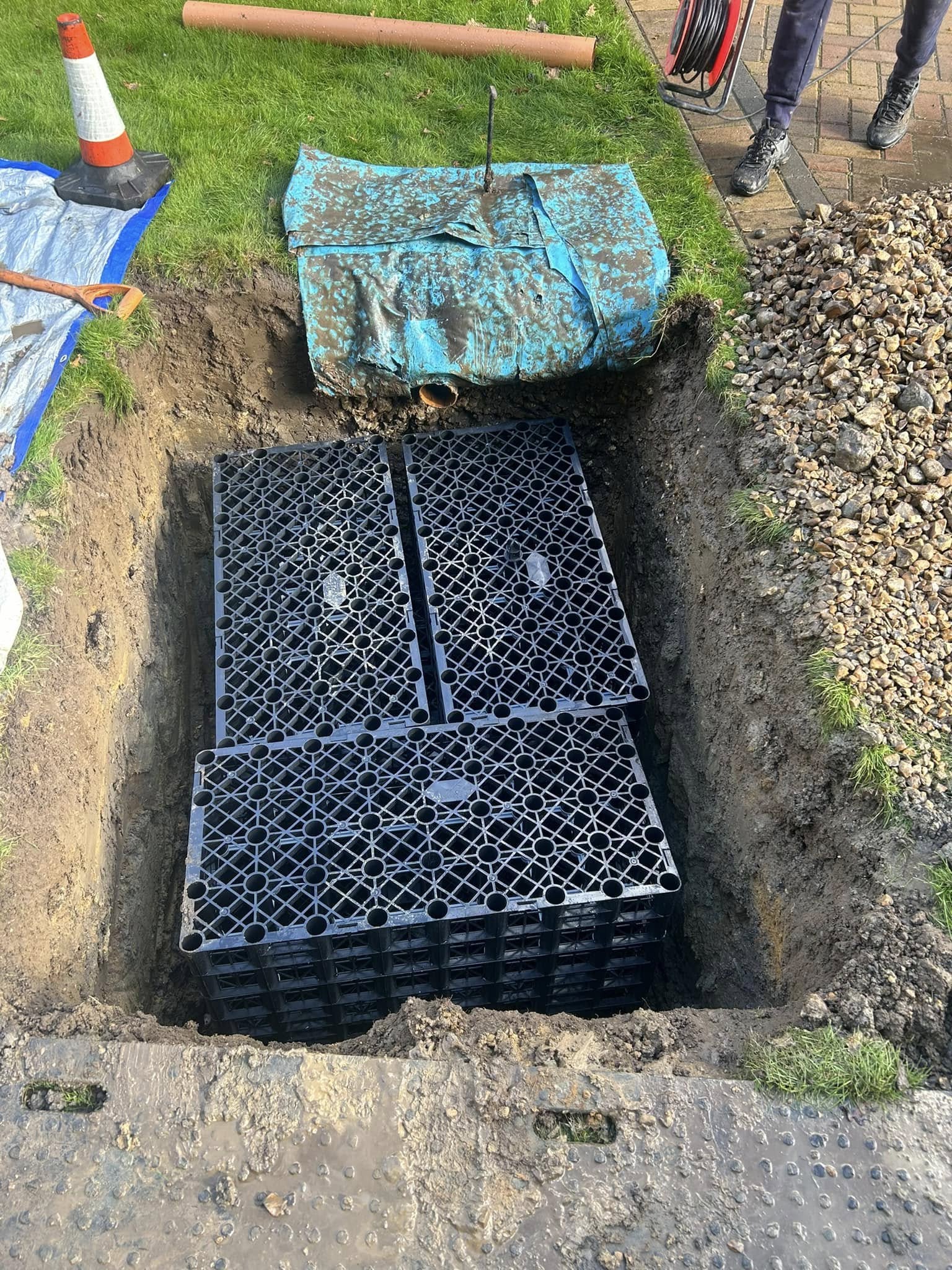 A rectangular pit dug in the ground contains stacked black plastic drainage crates. The surrounding area has grass, soil, stones, plastic sheeting, a traffic cone, and construction materials.