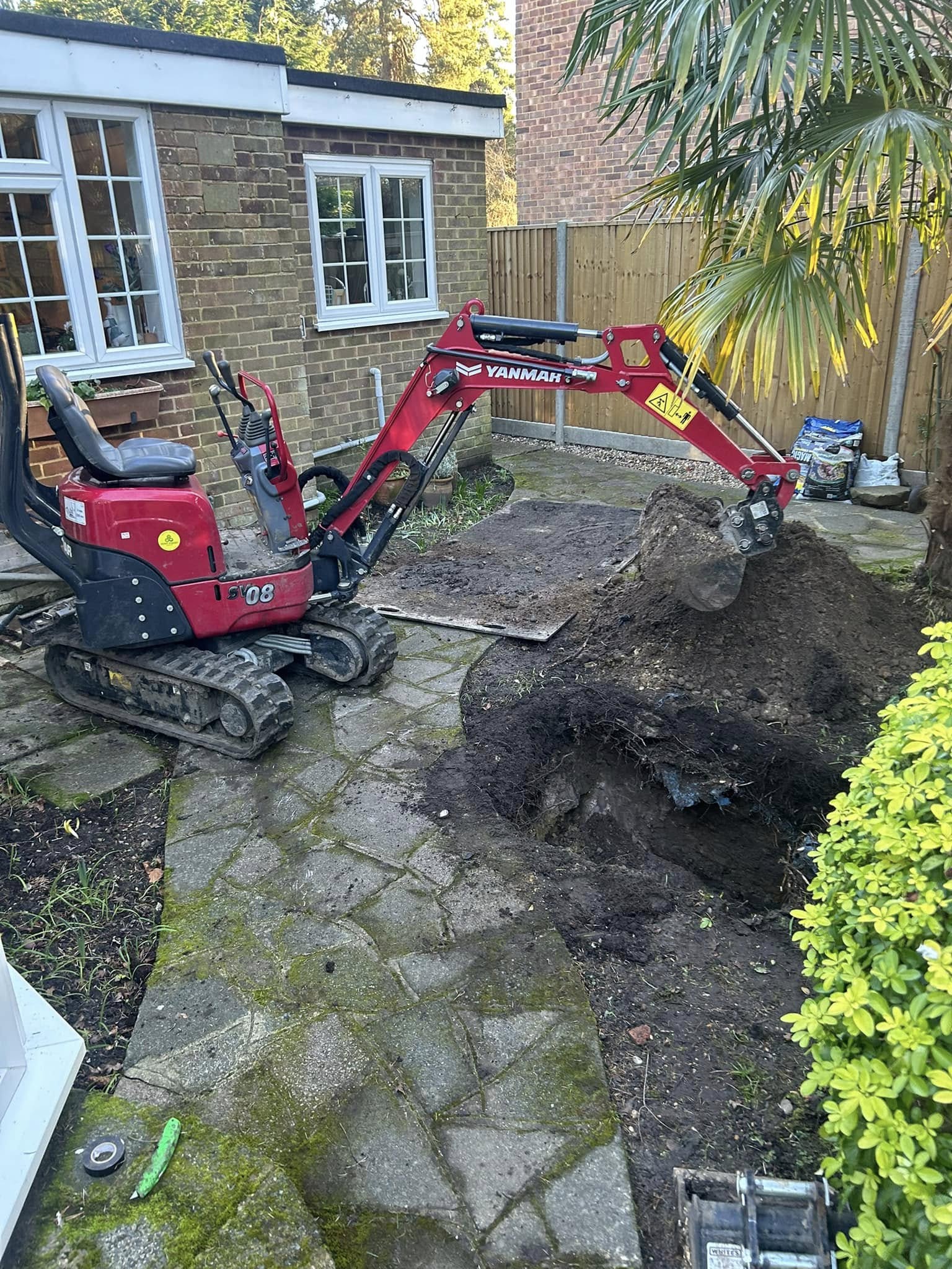 A small red excavator is digging up a large root ball in a garden next to a brick house. The yard has a stone path, green shrubs, and a wooden fence in the background.