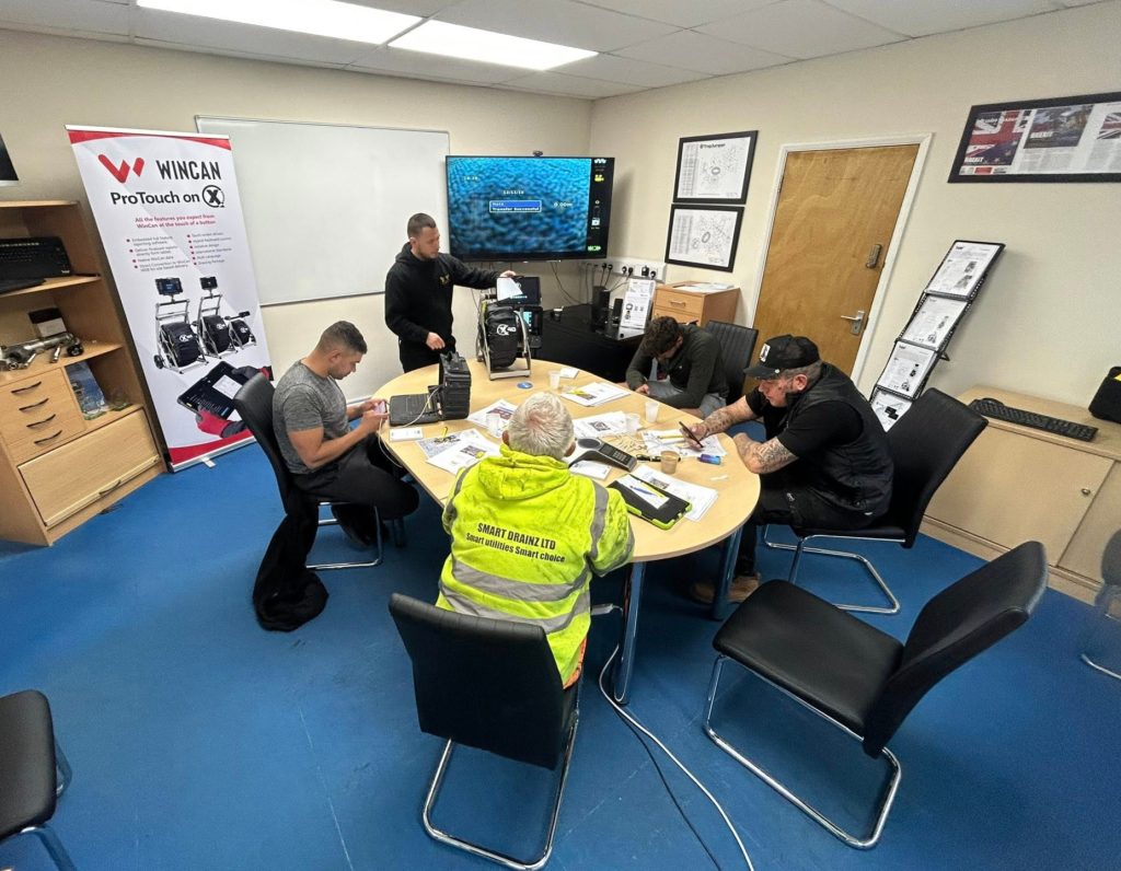 Five men are seated around a table in a meeting room with blue flooring, looking at papers and equipment. One man wears a bright yellow safety vest. Technical diagrams and a ProTouch On banner are visible in the background.