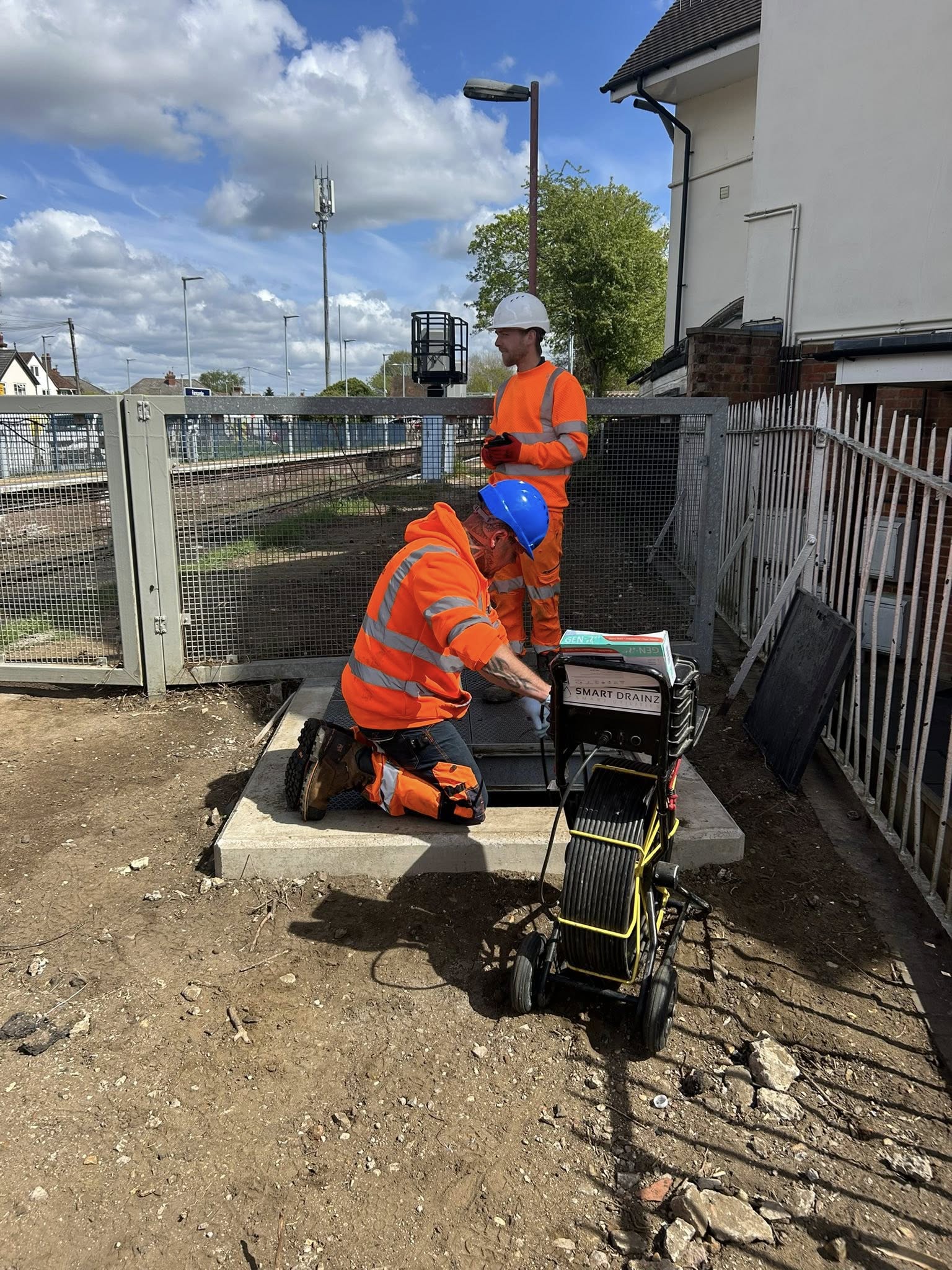 Two construction workers in orange safety gear and helmets work by a gated area near train tracks. One kneels over a utility box; the other stands nearby. Equipment and tools are set on a concrete pad.