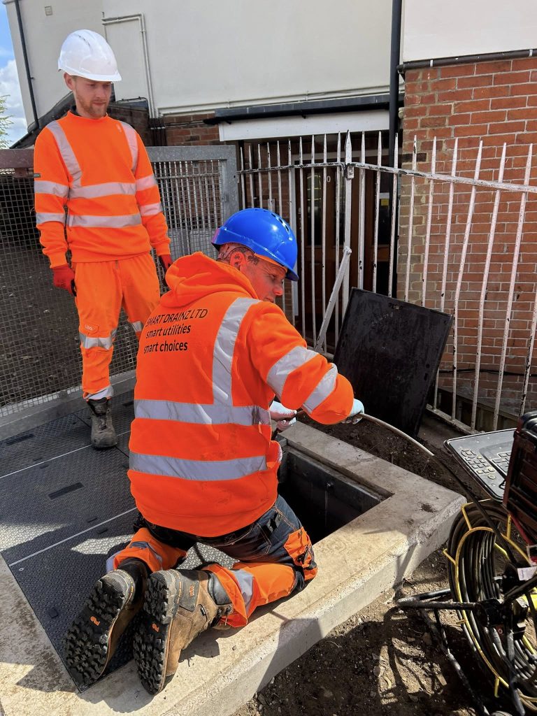 Two construction workers in orange high-visibility clothing and hard hats work beside an open underground access hatch. One is kneeling and working inside the hatch, while the other stands nearby watching.
